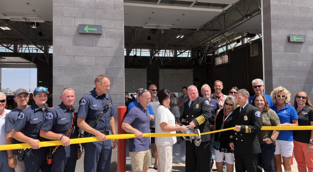 Mayor Randall and Fire Chief Stoker use a jaws of life tool to cut a fire hose as a symbol of a ribbon cutting. Related personnel are holding the house and posing behind them. 